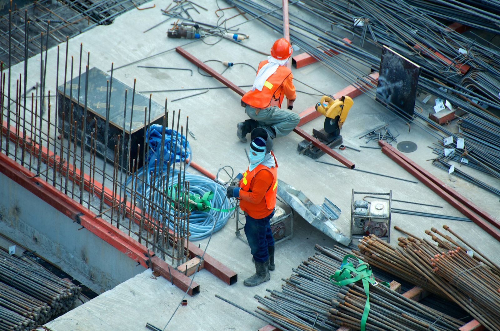Vue plongeante sur deux ouvriers en gilet orange au milieu des barres d'acier d'un chantier de gros œuvre.